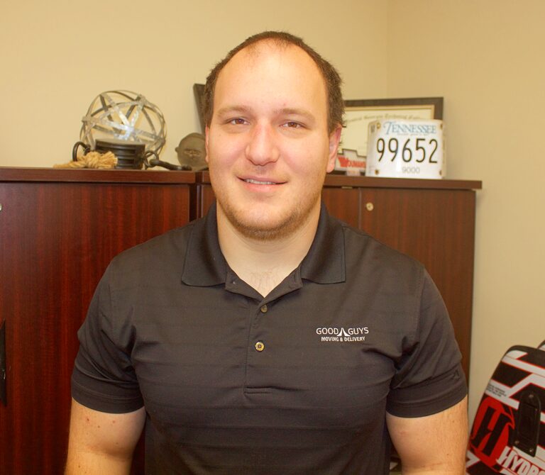 Smiling man in a black polo shirt sits indoors with a decorated background.